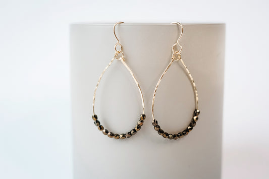 Gold teardrop shaped dangle earrings strung with 12 bronze-colored faceted glass beads that rest at the bottom of the teardrop shape, hanging from a beige color cylinder against a white background.
