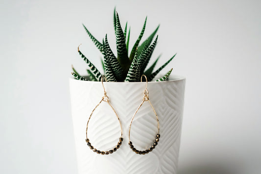 Gold teardrop shaped dangle earrings strung with 12 bronze-colored faceted glass beads that rest at the bottom of the teardrop shape, hanging from a white planter containing a small green plant.