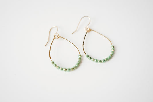 Gold teardrop shaped dangle earrings strung with 12 gold-highlighted green colored faceted glass beads that rest at the bottom of the teardrop shape, laying flat on a white background.