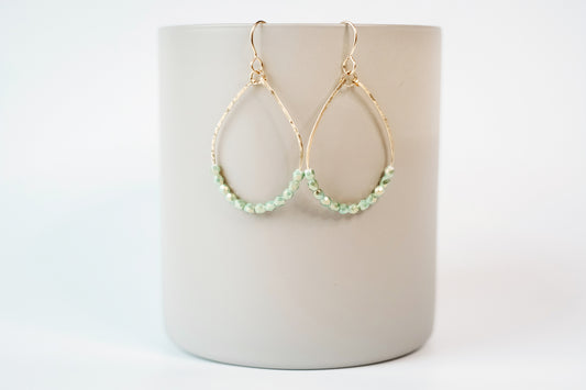 Gold teardrop shaped dangle earrings strung with 12 gold-highlighted green colored faceted glass beads that rest at the bottom of the teardrop shape, hanging from a beige cylinder on a white background.