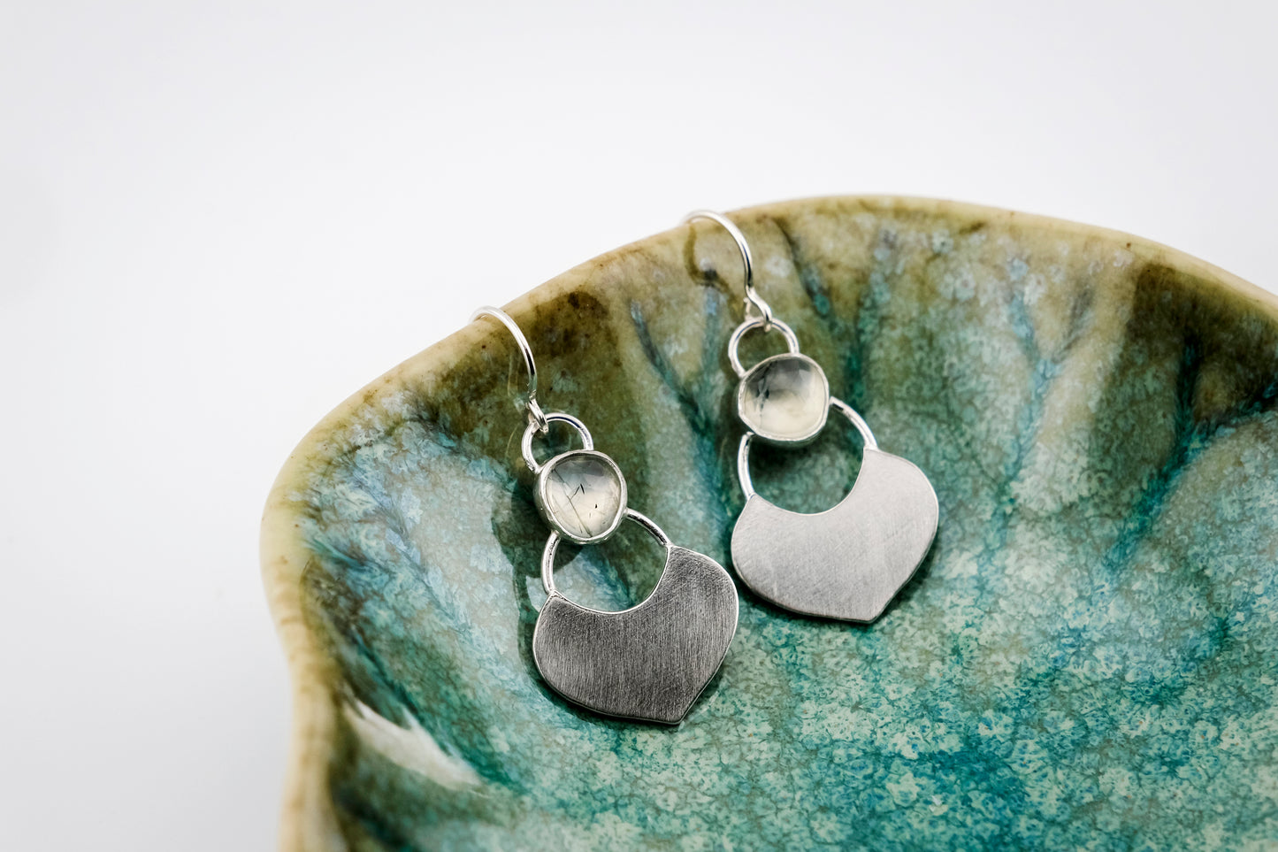 Silver dangle earrings featuring a prehnite green gemstone with a small flat shield-shaped silver dangle below, hanging from the edge of a small  green shallow dish against a white background.