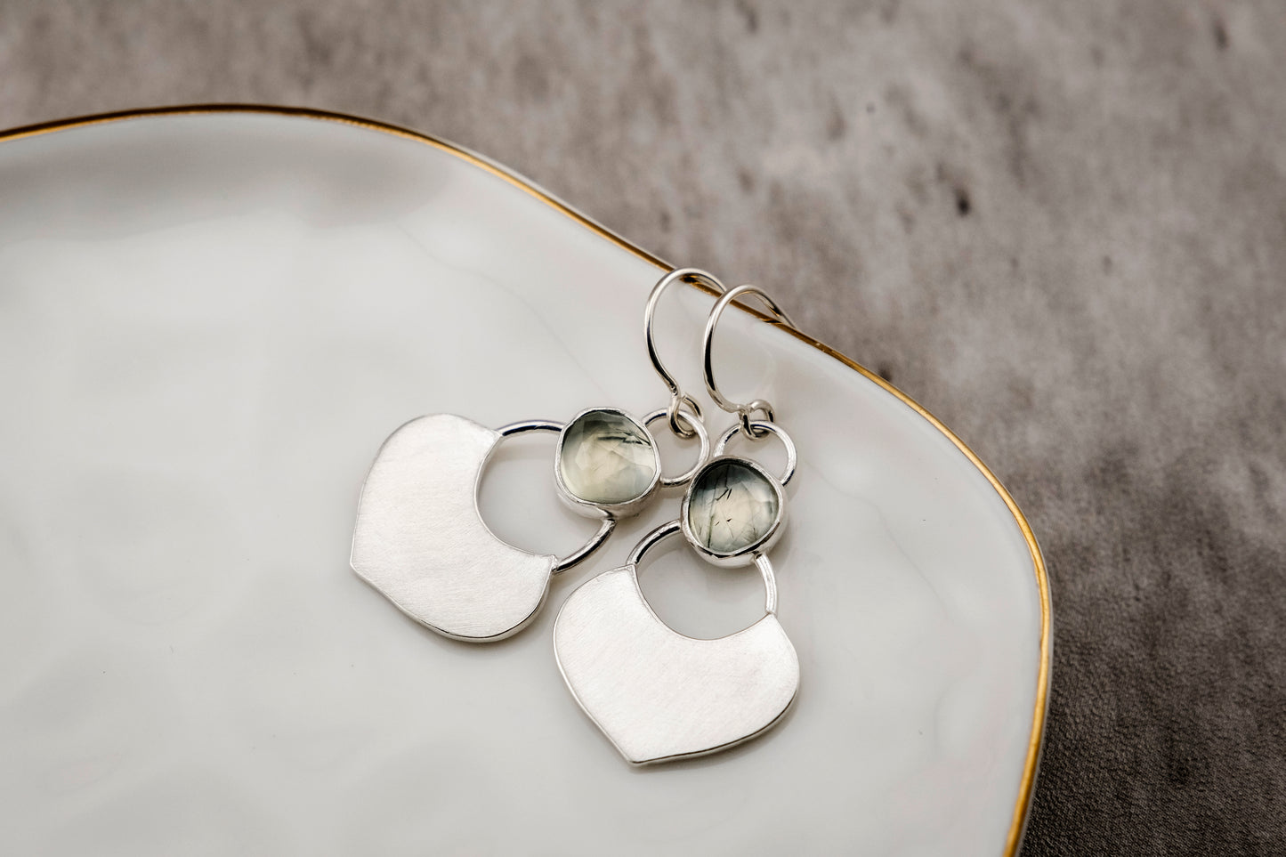 Silver dangle earrings featuring a prehnite green gemstone with a small flat shield-shaped silver dangle below, resting in a white shallow dish against a concrete surface.