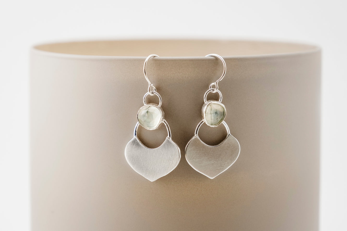 Silver dangle earrings featuring a prehnite green gemstone with a small flat shield-shaped silver dangle below, hanging from a beige cylinder against a white background.