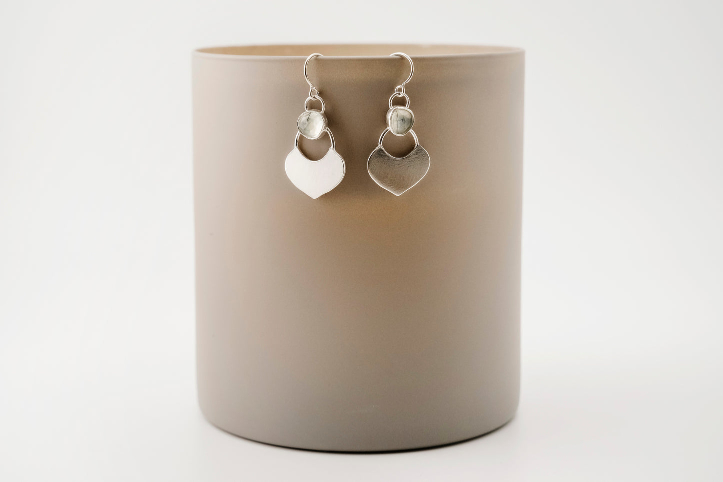 Silver dangle earrings featuring a prehnite green gemstone with a small flat shield-shaped silver dangle below, hanging from a beige cylinder against a white background.