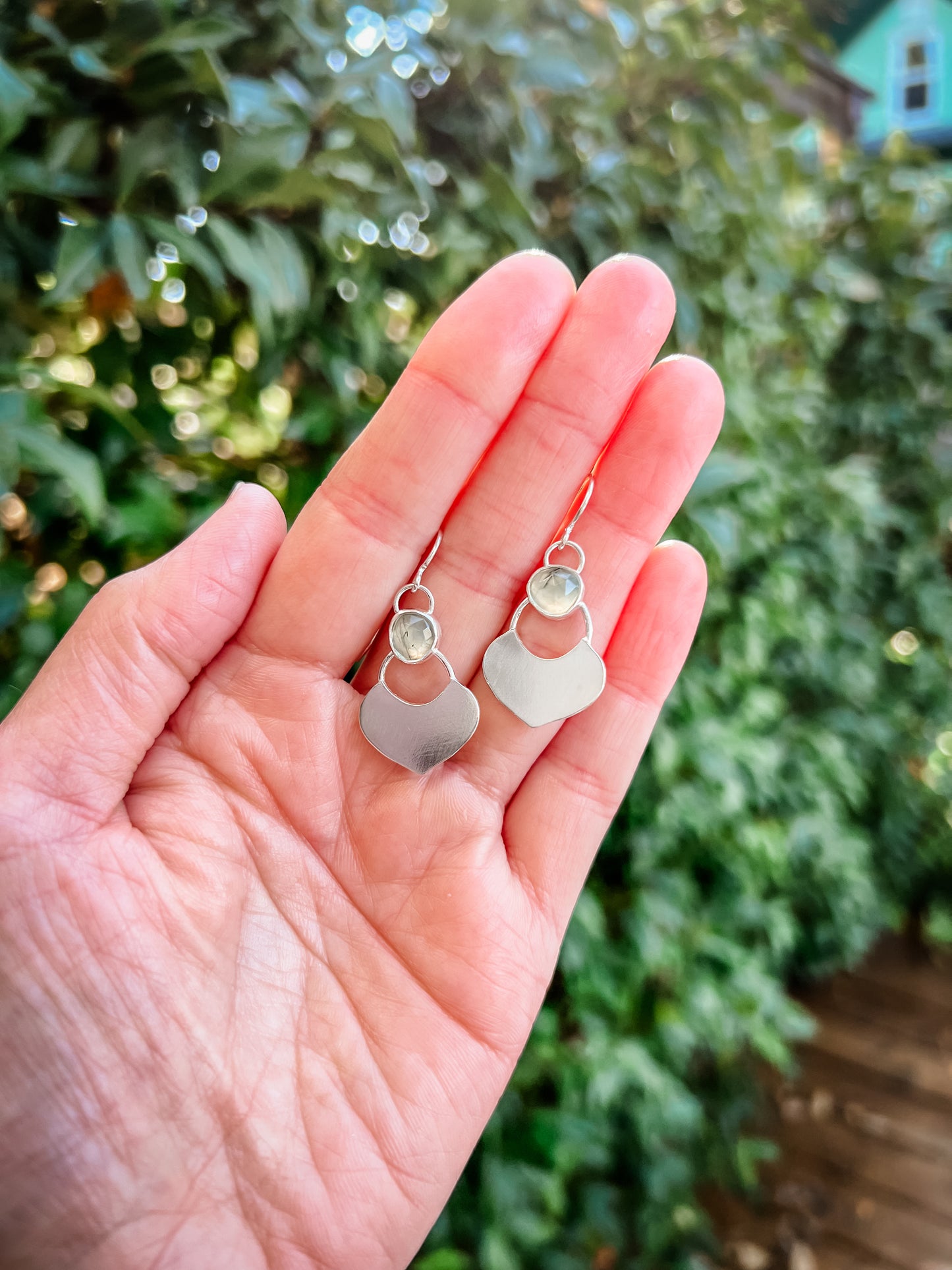 Silver dangle earrings featuring a prehnite green gemstone with a small flat shield-shaped silver dangle below, laying in the palm of the artist's hands.