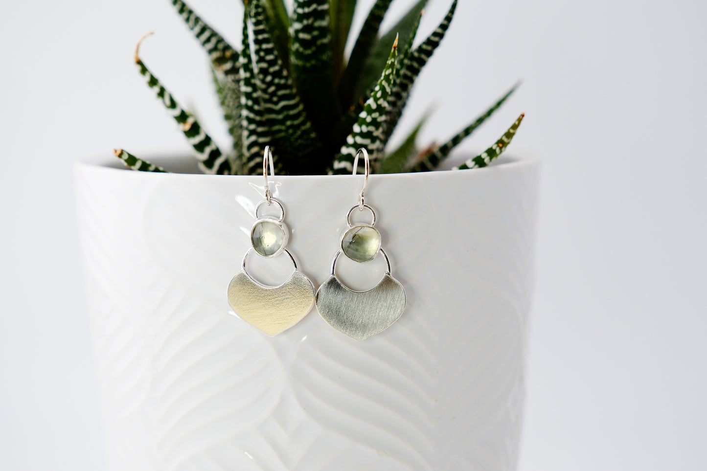 Silver dangle earrings featuring a prehnite green gemstone with a small flat shield-shaped silver dangle below, hanging from a small white plant pot which contains a green succulent.