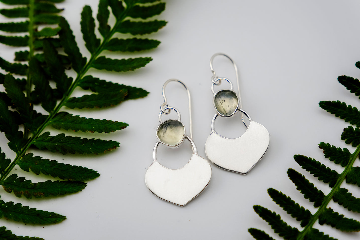 Silver dangle earrings featuring a prehnite green gemstone with a small flat shield-shaped silver dangle below, laying flat on a white surface.