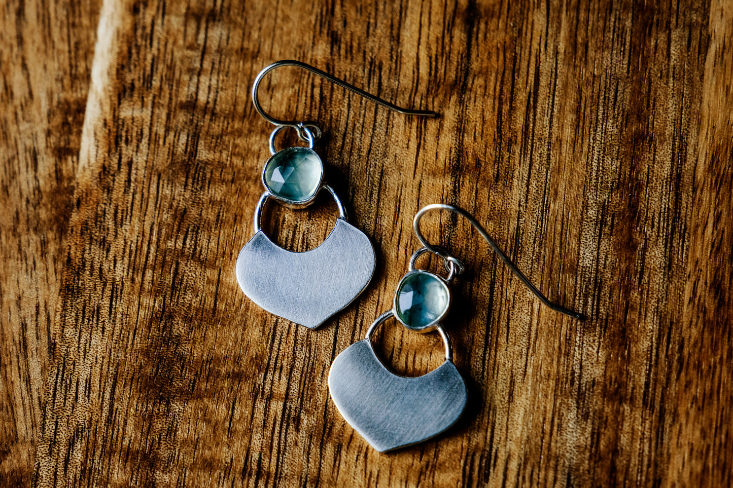 Silver dangle earrings featuring a prehnite green gemstone with a small flat shield-shaped silver dangle below,  laying flat on a wood surface.