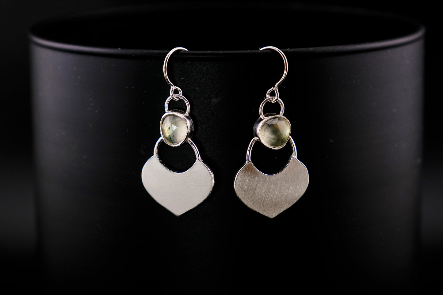 Silver dangle earrings featuring a prehnite green gemstone with a small flat shield-shaped silver dangle below, hanging from a black cylinder against a black background.