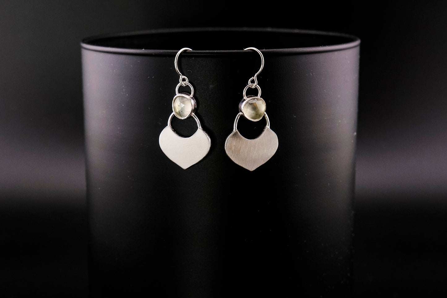 Silver dangle earrings featuring a prehnite green gemstone with a small flat shield-shaped silver dangle below, hanging from a black cylinder against a black background.