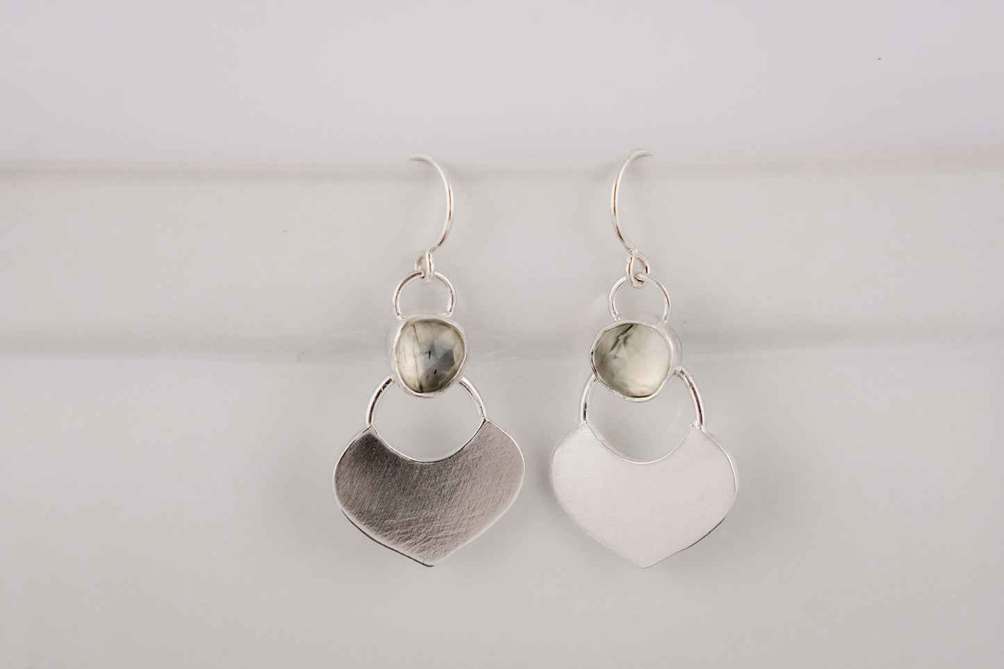 Silver dangle earrings featuring a prehnite green gemstone with a small flat shield-shaped silver dangle below, hanging  against a white background.