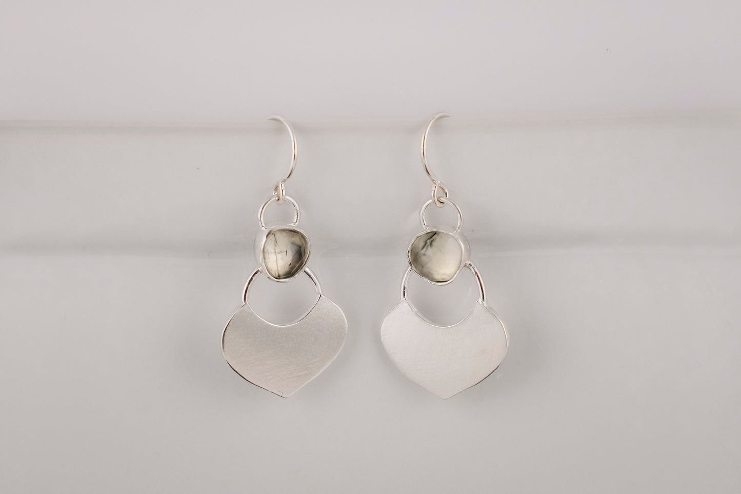 Silver dangle earrings featuring a prehnite green gemstone with a small flat shield-shaped silver dangle below, hanging  against a white background