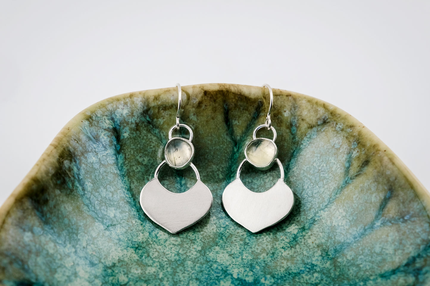 Silver dangle earrings featuring a prehnite green gemstone with a small flat shield-shaped silver dangle below, hanging from the edge of a small  green shallow dish against a white background.